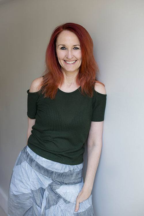 image of jill bennett wearing a green shirt and blue pale denim skirt she has red hair and is leaning against a wall