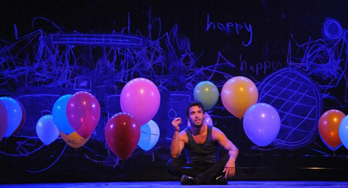 image of a man on stage chalk on wall behind says happy multi coloured balloons are behind him