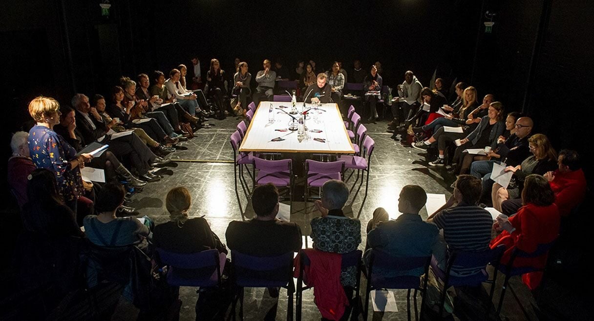 image of lois weaver and her long table. a table sits in the middle of the room with a circle of participants sitting around in chairs