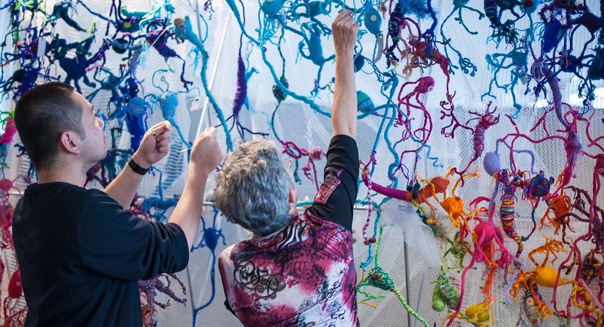 image of a man and woman standing in front a very large multicoloured wall hanging knitted artwork