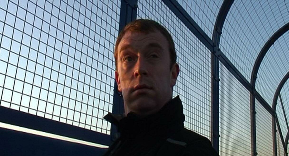 close up of a man's head he is looking sideways and a metal fence is behind him with blue sky