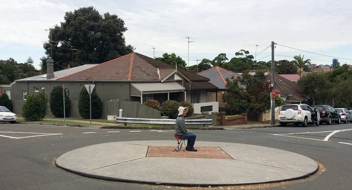 man is sitting on a stool in the middle of a suburban street in the centre of a roundabout