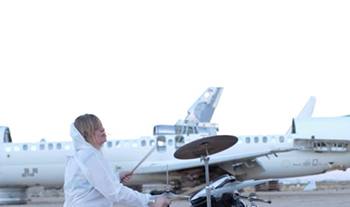 tina havelock stevens playing drums in a white suit in front of a plane