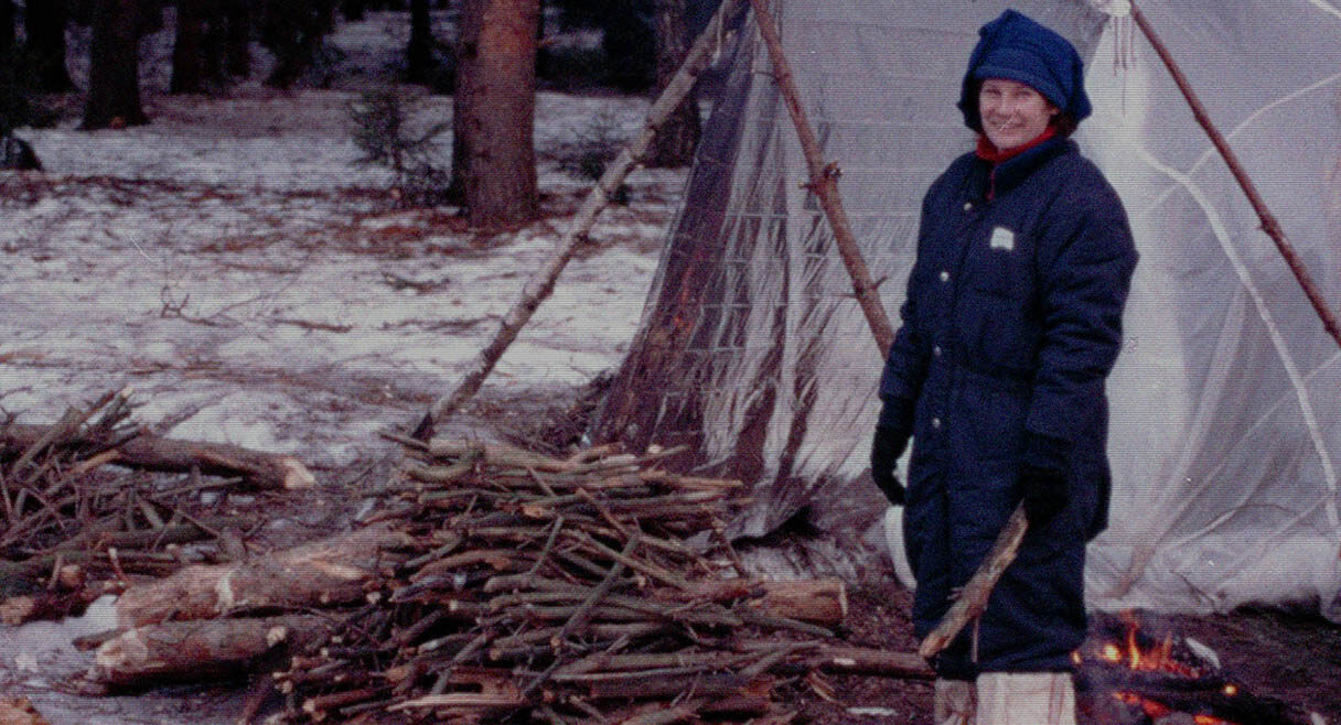 image of a woman standing in a forest with snow a white tent is beside her and she is surrounded by firewood