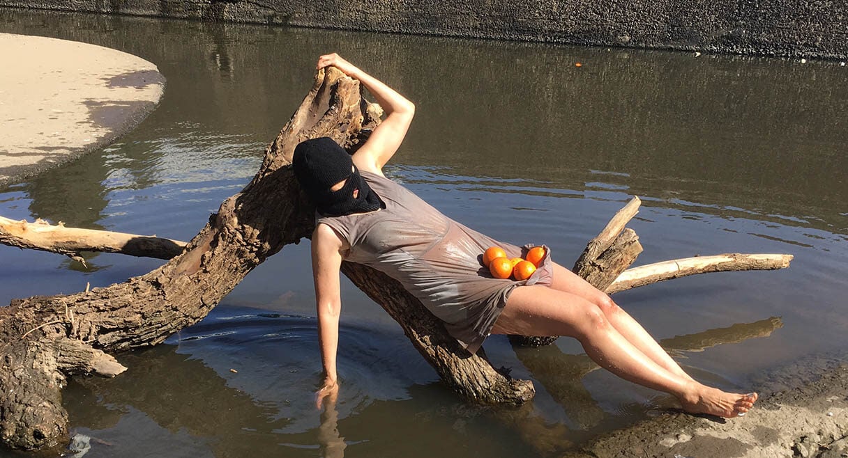 woman in a grey dress lies on a stump in a river she has a black mask with oranges in her lap