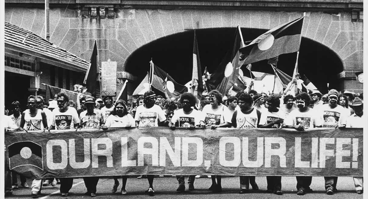 black and white photo of indigenous australians holding a banner 'our land our people'