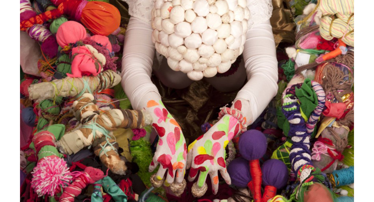 Image of figure laying face down into pile of multicoloured cloth with skin painted white with colourful shapes.